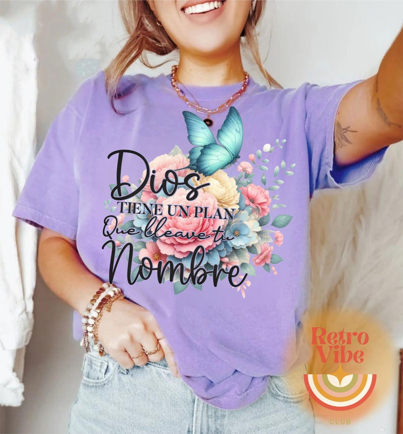 May include: A lilac-colored t-shirt featuring a floral design with a blue butterfly. The text "Dios tiene un plan que lleva tu nombre" is printed on the shirt. The design includes pink, yellow, and blue flowers.