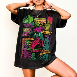 May include: Black t-shirt featuring a colorful graphic of classic Universal Monsters movie posters. The design includes images of Frankenstein, Dracula, the Mummy, and the Wolfman, with text like "Creature from the Black Lagoon."