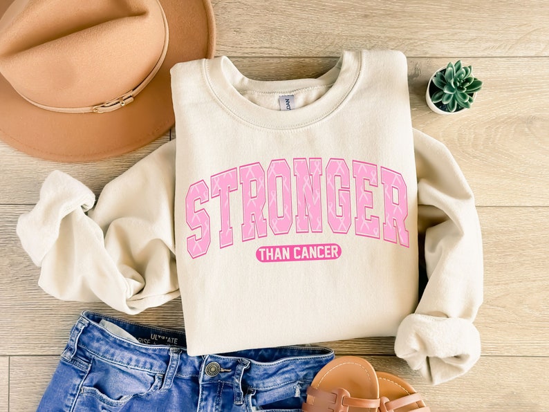 May include: Cream-colored sweatshirt with the text "STRONGER THAN CANCER" in pink, with pink ribbon accents. The sweatshirt is paired with blue jeans, a tan hat, and sandals. A succulent plant is also visible.