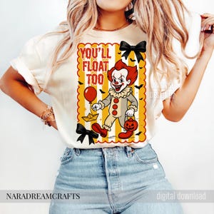 Op de afbeelding: Cr&egrave;mekleurig T-shirt met een cartoonclown met rood haar en make-up, die een ballon en een pompoen-lantaarn vasthoudt. De tekst "YOU'LL FLOAT TOO" staat boven de clown. Zwarte vleermuizen en strikken versieren het ontwerp, perfect voor Halloween.