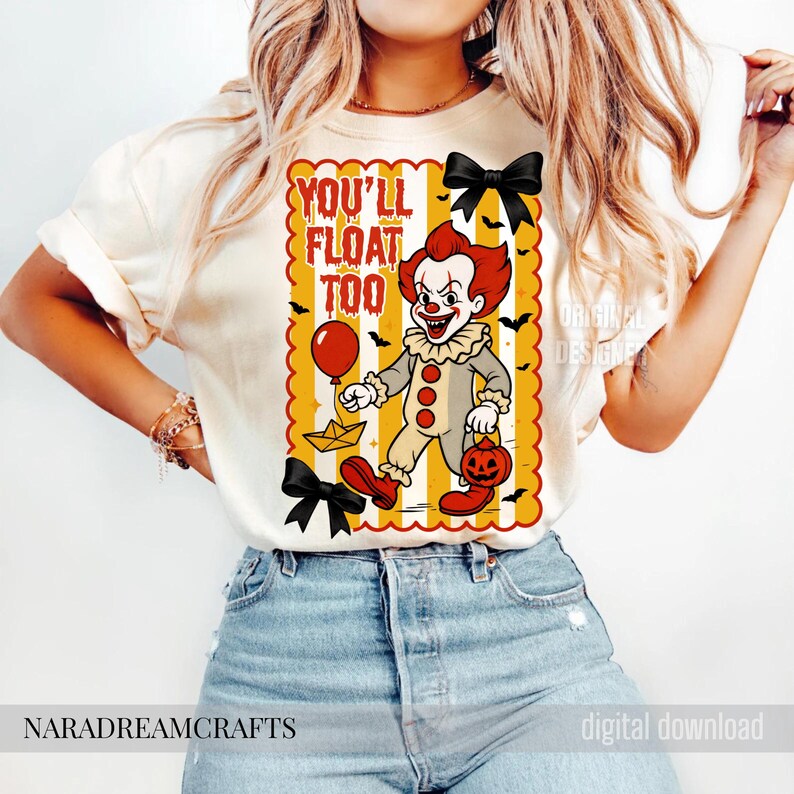 Op de afbeelding: Cr&egrave;mekleurig T-shirt met een cartoonclown met rood haar en make-up, die een ballon en een pompoen-lantaarn vasthoudt. De tekst "YOU'LL FLOAT TOO" staat boven de clown. Zwarte vleermuizen en strikken versieren het ontwerp, perfect voor Halloween.