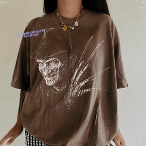 Puede incluir: Camiseta marr&oacute;n con un gr&aacute;fico blanco de Freddy Krueger. La imagen muestra un primer plano de la cara del personaje y el guante con cuchillas. El texto "NMADREAMCRAFTS do not copy" es visible en la camiseta.