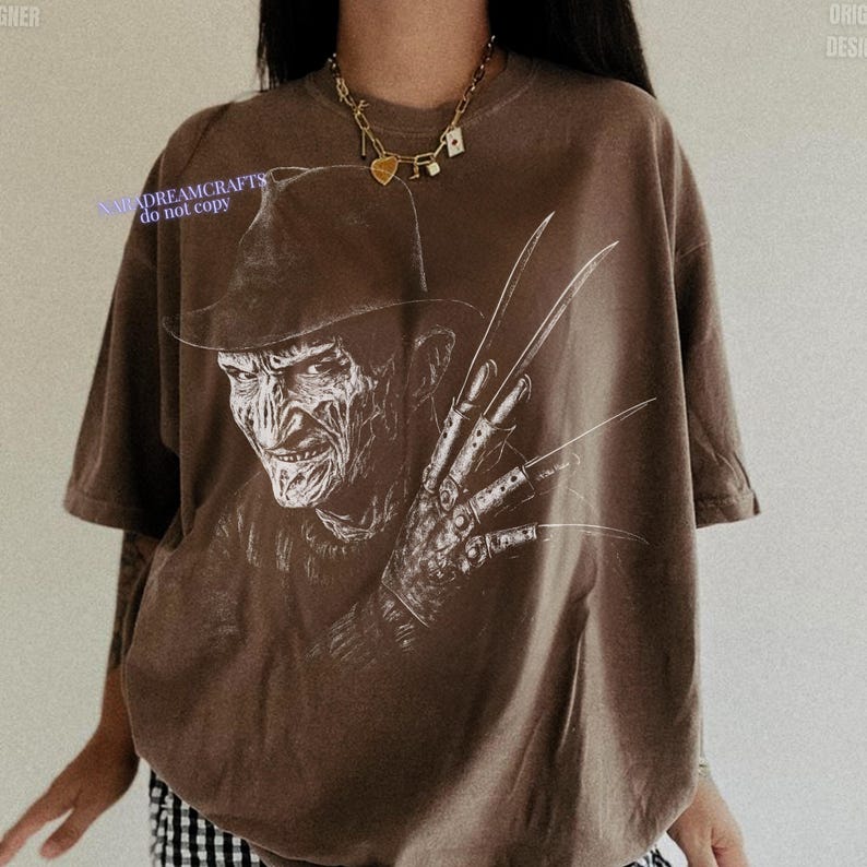 Puede incluir: Camiseta marr&oacute;n con un gr&aacute;fico blanco de Freddy Krueger. La imagen muestra un primer plano de la cara del personaje y el guante con cuchillas. El texto "NMADREAMCRAFTS do not copy" es visible en la camiseta.