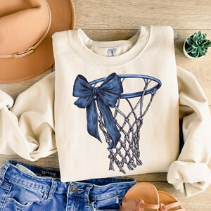 Peut inclure: Sweat-shirt cr&egrave;me avec un motif de panier de basket-ball bleu et un n&oelig;ud. Le sweat-shirt est associ&eacute; &agrave; un short en jean bleu, des sandales et un chapeau beige. Une petite plante succulente est &eacute;galement visible.