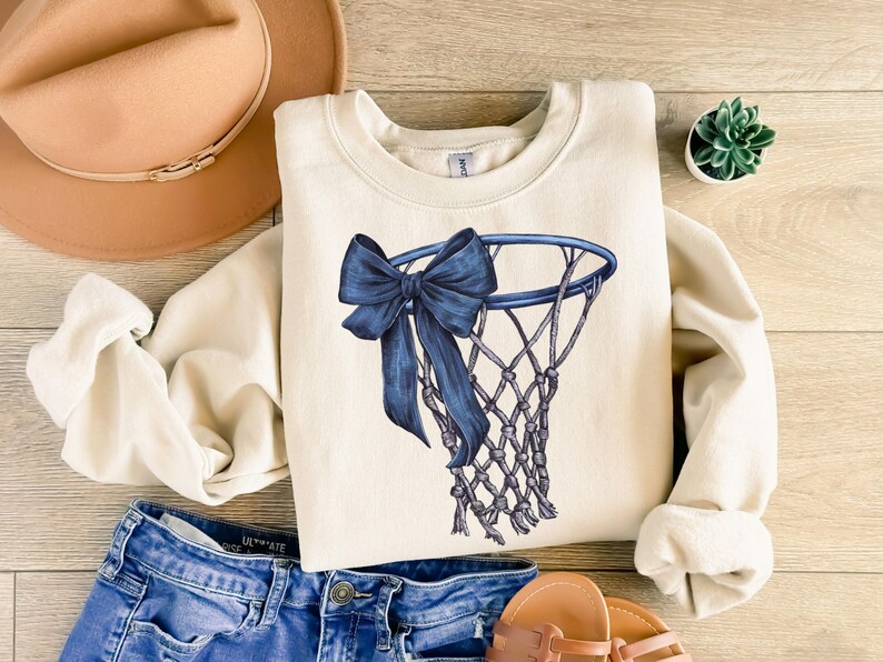 Peut inclure: Sweat-shirt cr&egrave;me avec un motif de panier de basket-ball bleu et un n&oelig;ud. Le sweat-shirt est associ&eacute; &agrave; un short en jean bleu, des sandales et un chapeau beige. Une petite plante succulente est &eacute;galement visible.