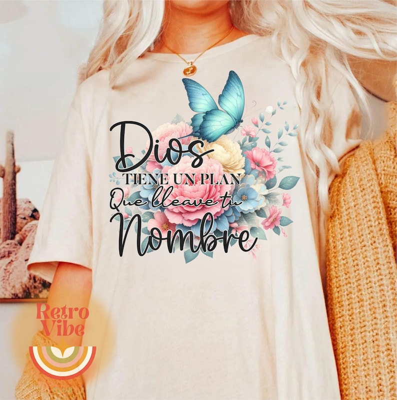 May include: Cream-colored t-shirt featuring a floral design with a blue butterfly and the Spanish text "Dios tiene un plan que lleve tu nombre." The design includes pink, blue, and yellow flowers. A retro rainbow logo is at the bottom.