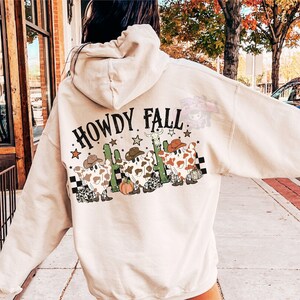 May include: Beige hoodie with the text "HOWDY FALL" and a graphic of three cow-shaped figures wearing cowboy hats, boots, and a cactus. The design includes stars and a checkered pattern. The hoodie has a drawstring hood.