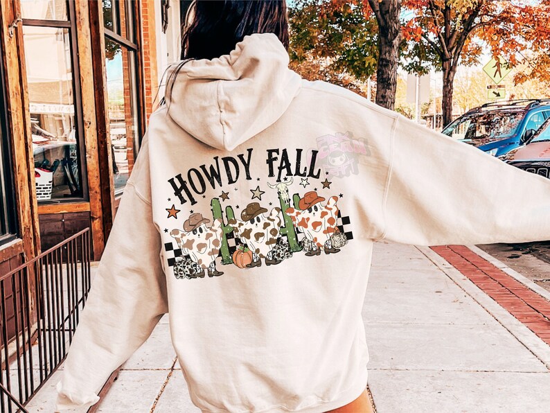 May include: Beige hoodie with the text "HOWDY FALL" and a graphic of three cow-shaped figures wearing cowboy hats, boots, and a cactus. The design includes stars and a checkered pattern. The hoodie has a drawstring hood.