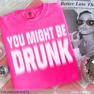 May include: A bright pink t-shirt with the text "YOU MIGHT BE DRUNK" in a layered white font. The shirt is a Comfort Colors brand. A black and white magazine clipping and a disco ball are in the background.