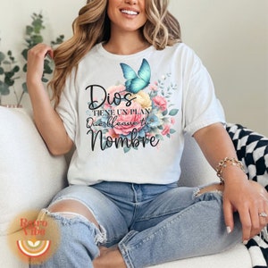 May include: White t-shirt featuring a floral design with a blue butterfly and the text "Dios tiene un plan Que lleve tu Nombre" in black. The design includes pink, blue, and yellow flowers. The shirt is worn with blue jeans.