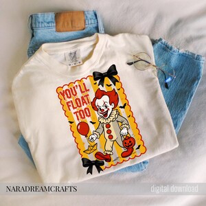 Op de afbeelding: Cr&egrave;mekleurig T-shirt met een cartoon clown afbeelding en de tekst "YOU'LL FLOAT TOO". De clown heeft rood haar, een witte gerimpelde kraag en houdt een rode ballon en een pompoen vast. Zwarte strikken en vleermuizen sieren het shirt.