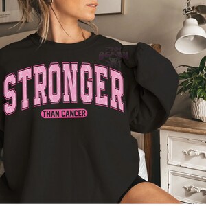 May include: Black sweatshirt with the words "STRONGER THAN CANCER" in pink block letters. The word "STRONGER" is arched across the top, and "THAN CANCER" is in a smaller, straight font below. The sweatshirt is a crew neck style.