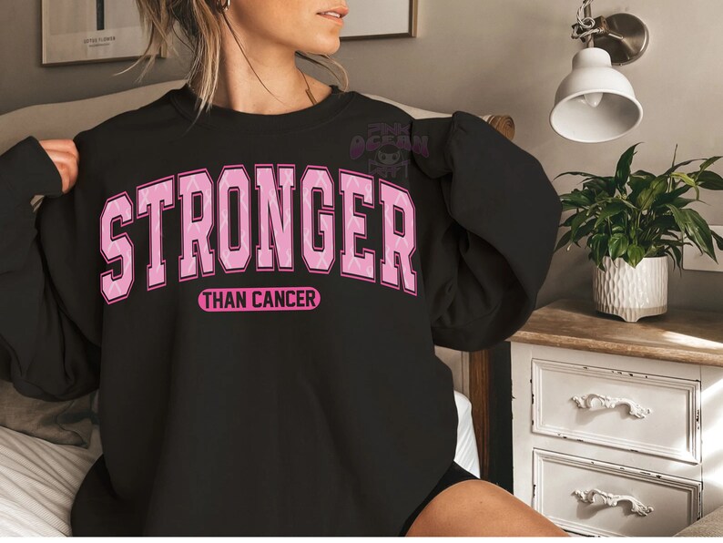 May include: Black sweatshirt with the words "STRONGER THAN CANCER" in pink block letters. The word "STRONGER" is arched across the top, and "THAN CANCER" is in a smaller, straight font below. The sweatshirt is a crew neck style.