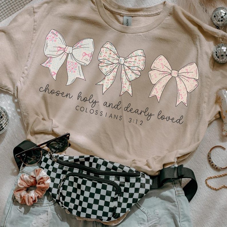 May include: A beige t-shirt featuring three decorative bows with floral patterns. The text "chosen, holy, and dearly loved" is printed below the bows, with "COLOSSIANS 3:12" underneath. A black and white checkered fanny pack, sunglasses, and a scrunchie are also visible.
