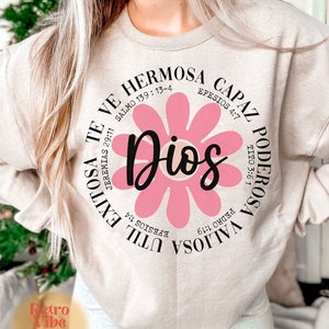 May include: A beige sweatshirt with a pink flower graphic and the text "Dios" in the center. The text around the flower includes bible verses in Spanish.