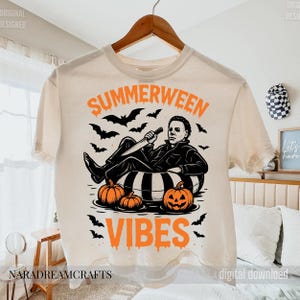 Op de afbeelding: Cr&egrave;mekleurig T-shirt met de oranje woorden "SUMMERWEEN VIBES", met een afbeelding van een horrorfilmkarakter dat ontspant in een zwemband met pompoenen en vleermuizen. Het shirt hangt aan een houten hanger.