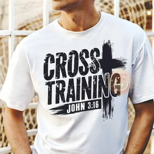 May include: White t-shirt with the words "CROSS TRAINING" in a distressed black font, accompanied by a black cross graphic. The text "JOHN 3:16" is below the main text. The shirt is a casual wear item.