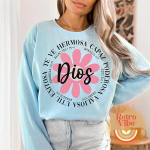 May include: Light blue sweatshirt with a pink flower graphic and the text "TE VE HERMOSA CAPAZ PODEROSA VALIOSA UTIL EXITOSA Dios" with bible verses surrounding the text.