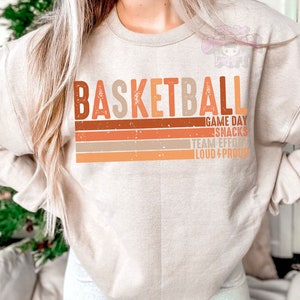 May include: A beige sweatshirt with the word "BASKETBALL" printed in orange, brown, and beige letters. The text "GAME DAY SNACKS TEAM EFFORT LOUD & PROUD" is printed below the word "BASKETBALL" in smaller letters.