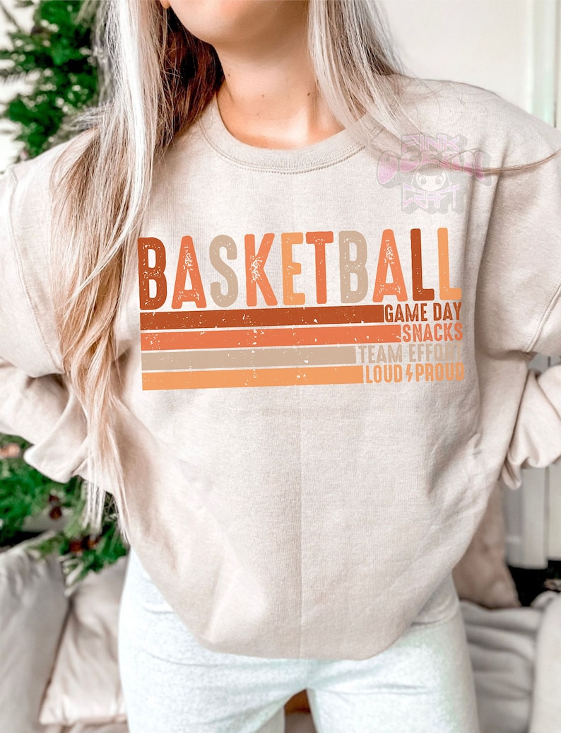 May include: A beige sweatshirt with the word "BASKETBALL" printed in orange, brown, and beige letters. The text "GAME DAY SNACKS TEAM EFFORT LOUD & PROUD" is printed below the word "BASKETBALL" in smaller letters.