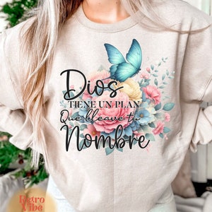 May include: Beige sweatshirt featuring a floral design with a blue butterfly and the Spanish text "Dios Tiene Un Plan Que Lleva Tu Nombre." The design includes pink, blue, and yellow flowers. The Retro Vibe Club logo is at the bottom.
