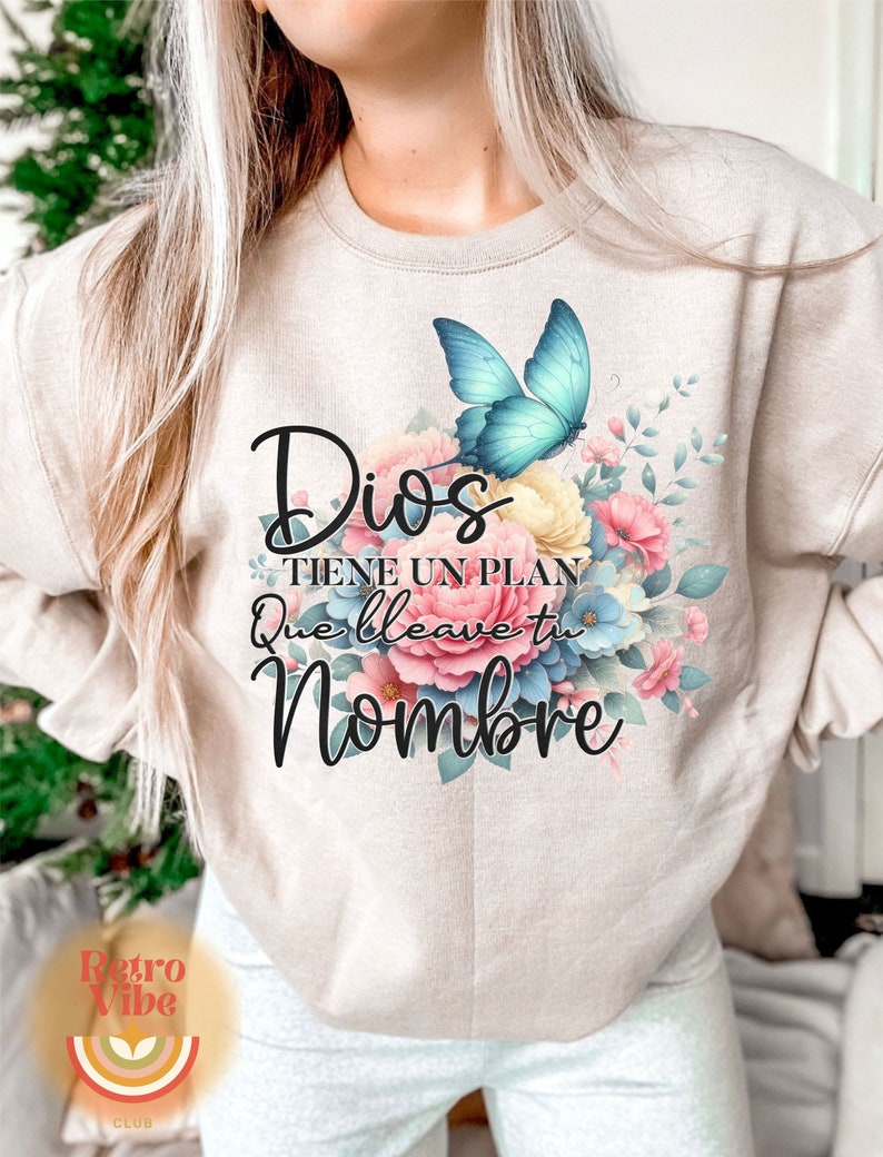 May include: Beige sweatshirt featuring a floral design with a blue butterfly and the Spanish text "Dios Tiene Un Plan Que Lleva Tu Nombre." The design includes pink, blue, and yellow flowers. The Retro Vibe Club logo is at the bottom.