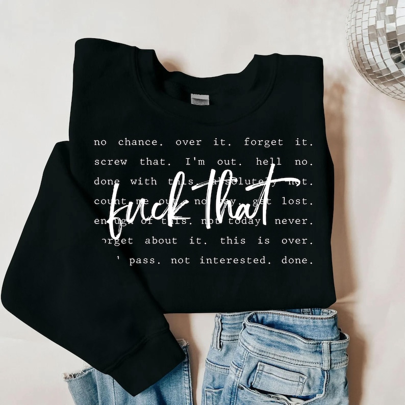 May include: Black sweatshirt with the phrase "fuck that" in white script. Additional text in white includes phrases like "no chance," "forget it," and "not interested." The sweatshirt is paired with blue jeans.