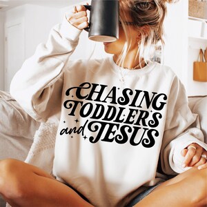 May include: White sweatshirt with black text that reads "Chasing Toddlers and Jesus" in a retro font.