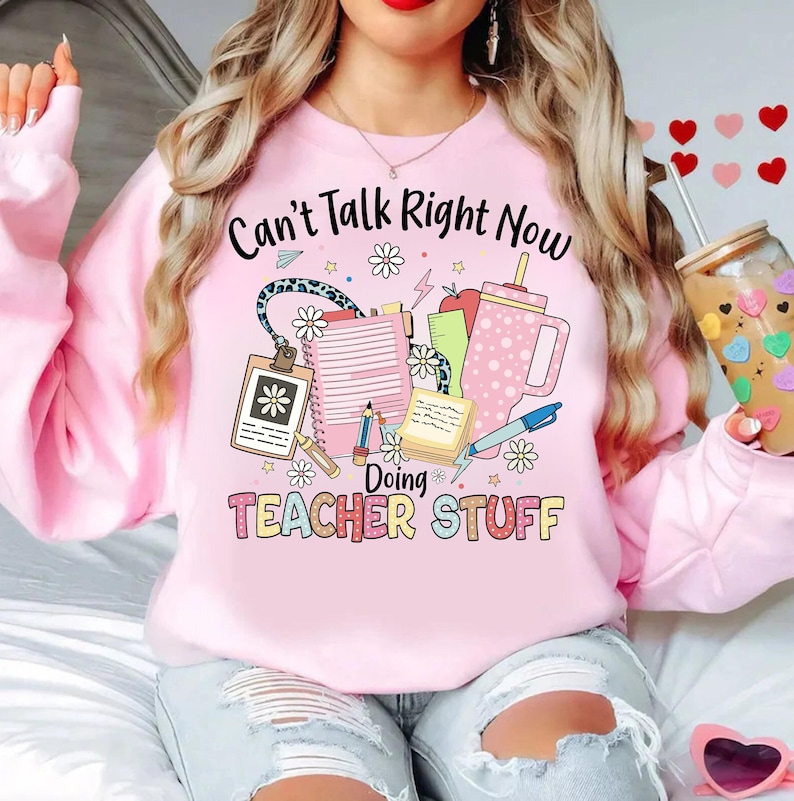 May include: Pink sweatshirt with the text "Can't Talk Right Now Doing Teacher Stuff" and a graphic of school supplies, including a pencil, a ruler, a notebook, a coffee cup, and flowers.