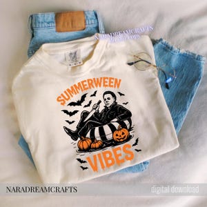 Op de afbeelding: Cr&egrave;mekleurige sweatshirt met de woorden "SUMMERWEEN VIBES" in oranje, met een afbeelding van een personage met een mes, pompoenen en vleermuizen. Het shirt ligt op een blauwe jeans.