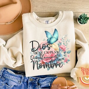 May include: Cream-colored sweatshirt with a floral and butterfly design. The design includes a blue butterfly and the Spanish text "Dios tiene un plan que lleve tu nombre". The sweatshirt is paired with blue jeans and sandals.