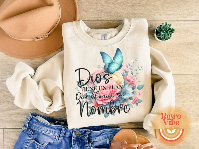 May include: Cream-colored sweatshirt with a floral and butterfly design. The design includes a blue butterfly and the Spanish text "Dios tiene un plan que lleve tu nombre". The sweatshirt is paired with blue jeans and sandals.