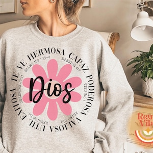 May include: A gray sweatshirt with a pink flower graphic and the text "Dios" in the center. The flower is surrounded by a circle of text in Spanish, including Bible verses.
