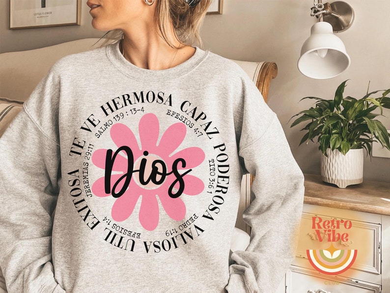 May include: A gray sweatshirt with a pink flower graphic and the text "Dios" in the center. The flower is surrounded by a circle of text in Spanish, including Bible verses.