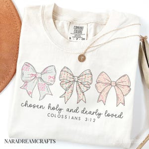 May include: Cream-colored t-shirt featuring three floral bows in pink and green. The text "chosen, holy, and dearly loved" is printed below the bows, with "COLOSSIANS 3:12" underneath. A gold necklace with a pendant is draped on the shirt.