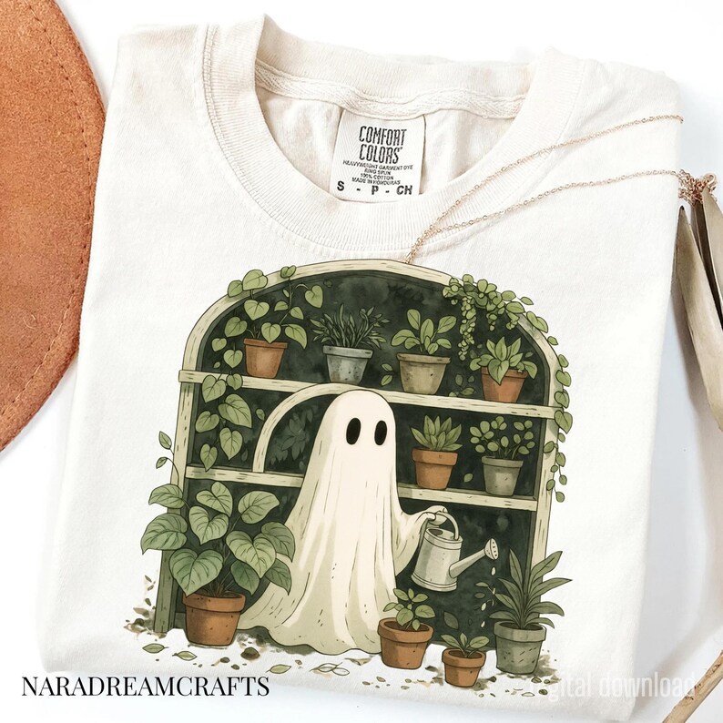 K&ouml;nnte beinhalten: Cremefarbenes T-Shirt mit einer skurrilen Illustration eines Geistes, der Pflanzen in einem Gew&auml;chshaus gie&szlig;t. Das Design umfasst verschiedene Topfpflanzen und den Text "NARADREAMCRAFTS". Das Shirt hat ein Comfort Colors-Etikett.