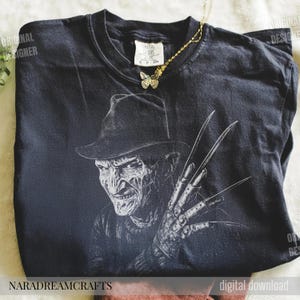 Puede incluir: Camiseta negra con un gr&aacute;fico de Freddy Krueger con sombrero y guante con garras. Un collar con colgante de mariposa dorada descansa sobre la camiseta. La camiseta tiene las palabras "Original Designer" y "NARADREAMCRAFTS".