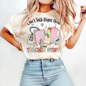 May include: A white t-shirt with the text "Can't Talk Right Now Doing Teacher Stuff" in pink and black lettering. The text is surrounded by colorful illustrations of school supplies, including a pencil, a ruler, a notebook, a clipboard, and a coffee cup.