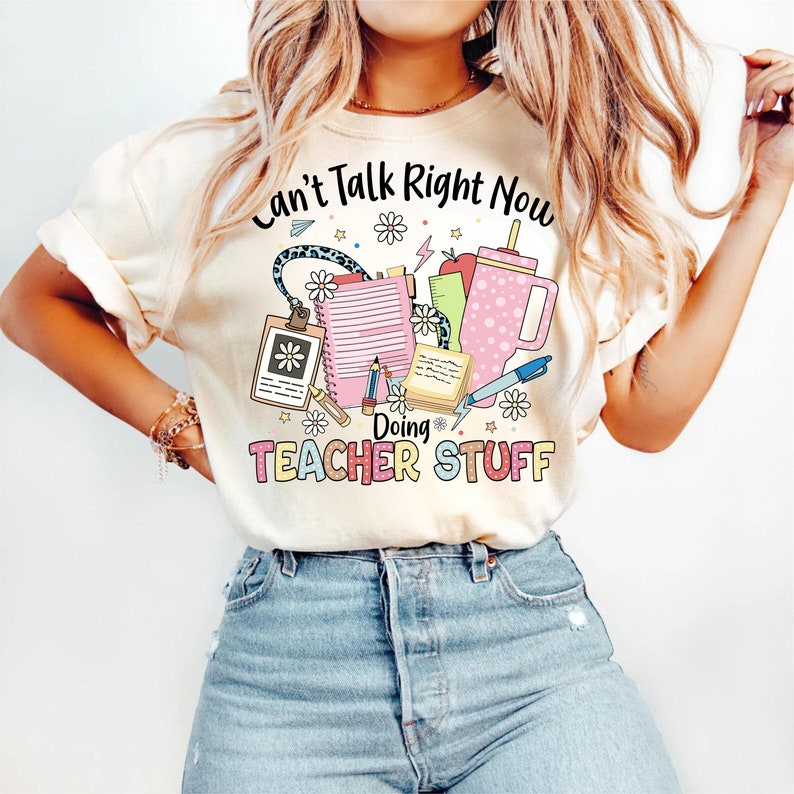 May include: A white t-shirt with the text "Can't Talk Right Now Doing Teacher Stuff" in pink and black lettering. The text is surrounded by colorful illustrations of school supplies, including a pencil, a ruler, a notebook, a clipboard, and a coffee cup.