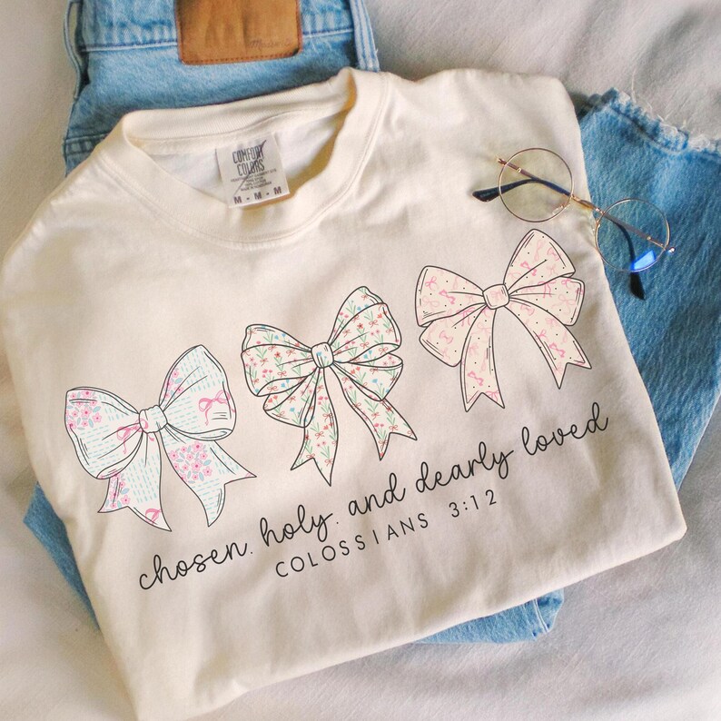 May include: Cream-colored t-shirt with three floral bow designs and the text "chosen, holy, and dearly loved" with "COLOSSIANS 3:12" below. The shirt is laid on top of blue jeans.