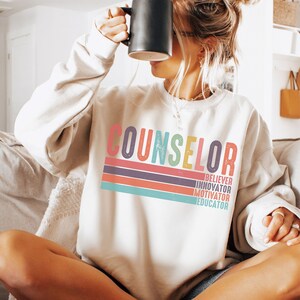 May include: White sweatshirt with the word "COUNSELOR" in rainbow colors and the words "BELIEVER, INNOVATOR, MOTIVATOR, EDUCATOR" underneath in black.