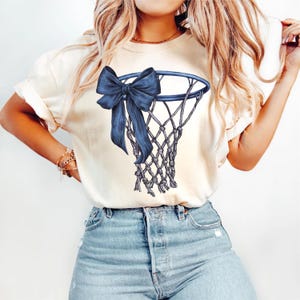 Peut inclure: T-shirt cr&egrave;me avec un motif de filet de basket-ball bleu marine orn&eacute; d'un grand n&oelig;ud. Le filet est d&eacute;taill&eacute; avec un motif tiss&eacute;, et le n&oelig;ud ajoute un &eacute;l&eacute;ment d&eacute;coratif. Le t-shirt est associ&eacute; &agrave; un jean en denim bleu clair.