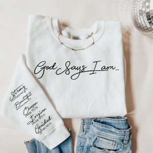 Może przedstawiać: Biała bluza z nadrukiem "God Says I am..." na przodzie. Lewy rękaw ma nadrukowane słowa "Strong, Beautiful, Chosen, Forgiven, Guided" wraz z odpowiadającymi im wersetami biblijnymi.
