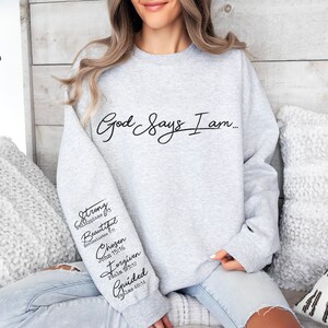 Może przedstawiać: Szara bluza z nadrukiem "God Says I am..." na przodzie. Lewy rękaw ma pionowo nadrukowane słowa "Strong, Beautiful, Chosen, Forgiven, Guided" wraz z odpowiadającymi im wersetami biblijnymi.