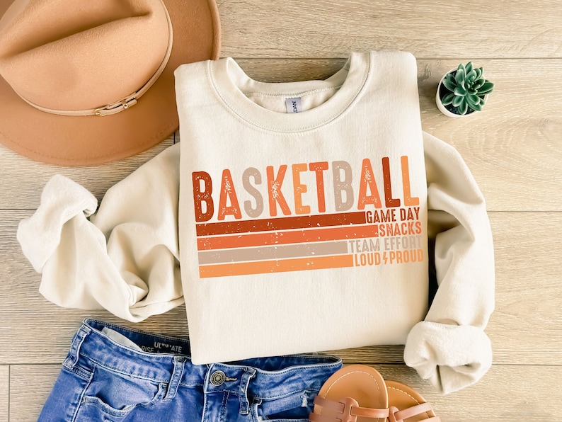 May include: A cream-colored sweatshirt with the word "BASKETBALL" in orange, red, and brown letters. The letters are layered over a retro-style striped design in orange, brown, and beige. The text "GAME DAY SNACKS TEAM EFFORT LOUD & PROUD" is printed in smaller letters below the word "BASKETBALL".
