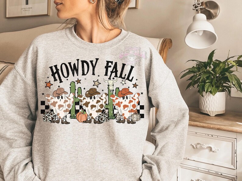 May include: A light gray sweatshirt with the words "HOWDY FALL" in black. The graphic features cartoon ghosts wearing cowboy hats and cowhide patterns, with cacti, stars, and a pumpkin.
