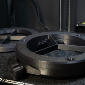 May include: Two black, circular 3D-printed objects with a cross-shaped interior design. The objects are on a textured black surface, possibly a 3D printer bed. One object has a knob attached. The objects are likely components for a larger device.