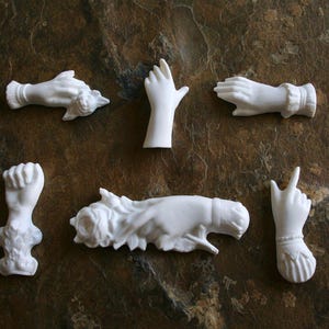 Pode incluir: Conjunto de seis estatuetas de mão de porcelana branca. As mãos estão em várias poses, incluindo segurando uma flor, apontando e fazendo um punho.