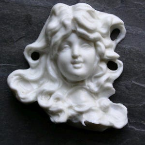 May include: A white, sculpted cameo of a woman's face with flowing hair. The detailed carving features a serene expression and is set against a dark, textured background. The cameo has a classical, artistic theme.