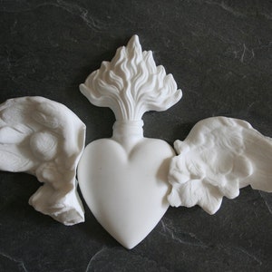 May include: A white matte ceramic wall ornament shaped like a central heart with a flame rising from the top and two detailed wings extending from the sides. The piece features a smooth finish on the heart and textured, sculpted feathers on the wings.